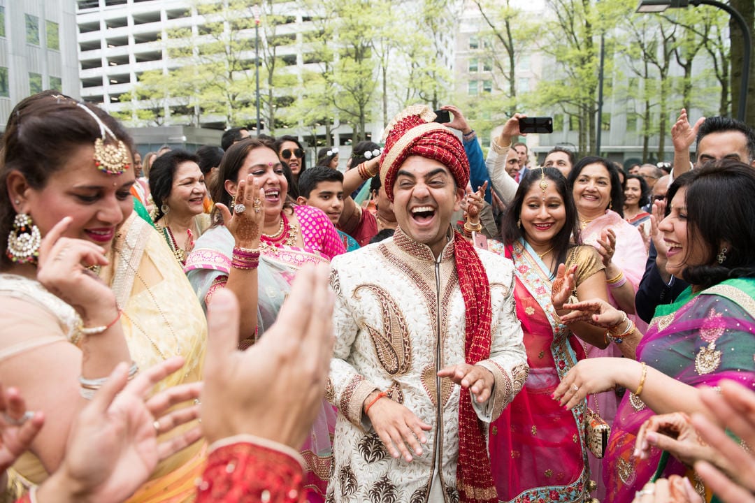 A groom laughs as he dances outdoors with women in traditional Indian garb.