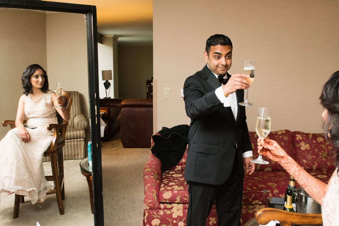 A groom wearing a black tuxedo toasts his bride in a long dress, reflected in a floor-length mirror.
