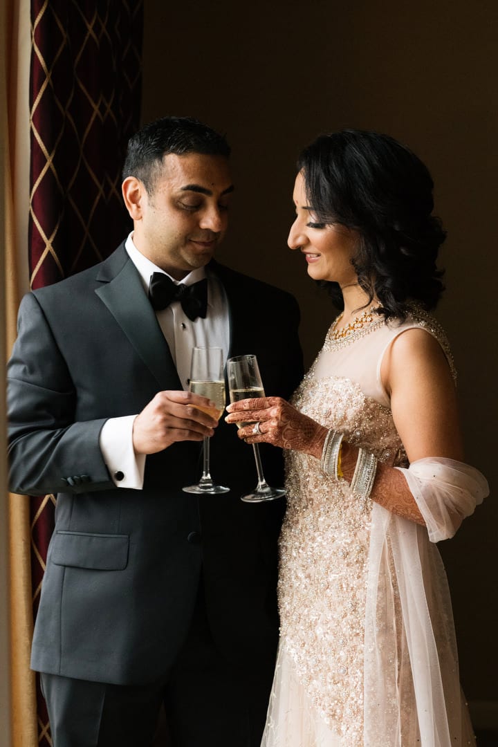 A bride and groom wearing formal evening attire toast each other with champagne.