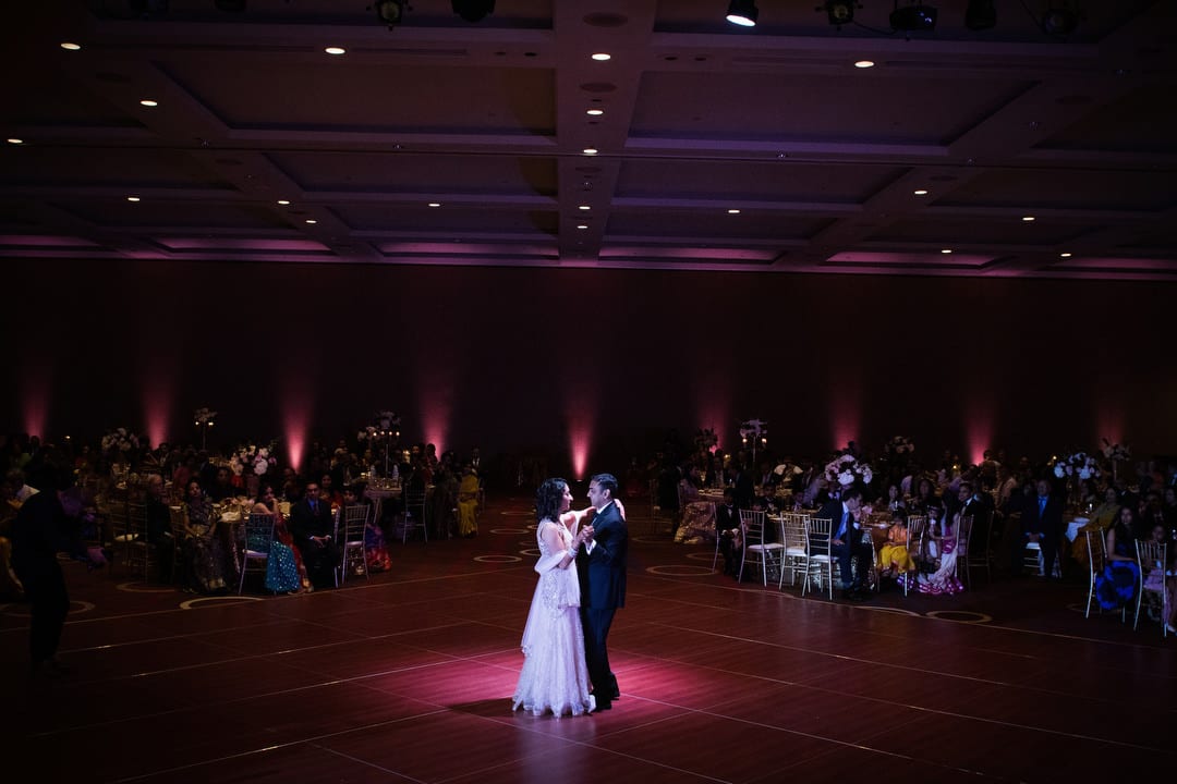 Lit by a single spot, a bride and groom share a first dance in a darkened ballroom.