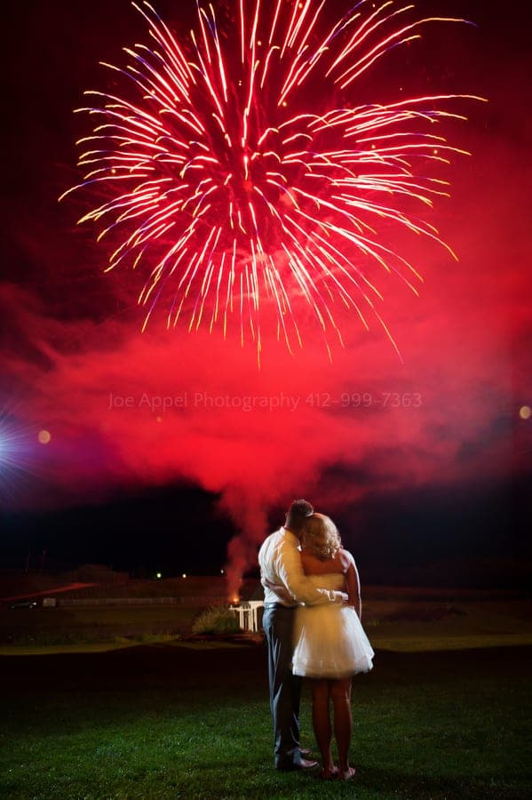 red fireworks explode above a bride and groom as they embrace at the end of their wedding reception