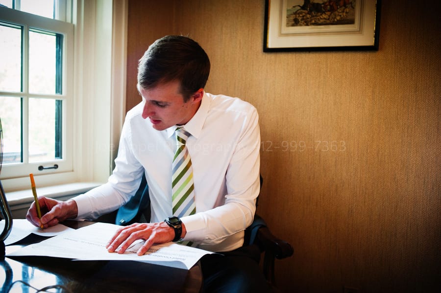 A groom quietly writes his wedding vows while seated next to a window.