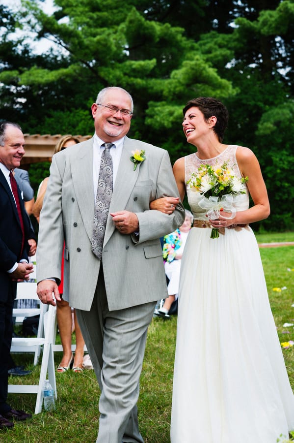 A bride and her father laugh as they walk down the aisle together at her wedding.
