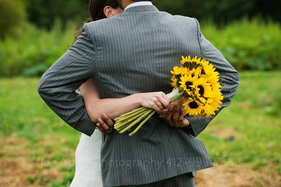 a photo from behind the groom as his bride hugs him while holding a bouquet of sunflowers outdoor wedding in pittsburgh