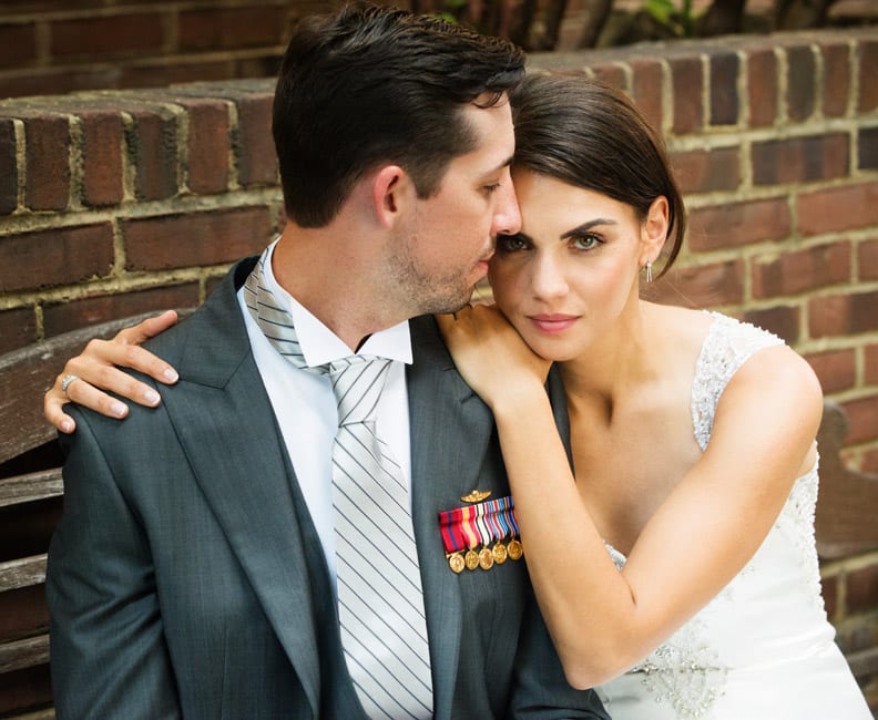 A bride holds her arms around the shoulders of her groom as they sit on a bench behind Cafe Milano in Georgetown. He has a gray tuxedo jacket with military medals.