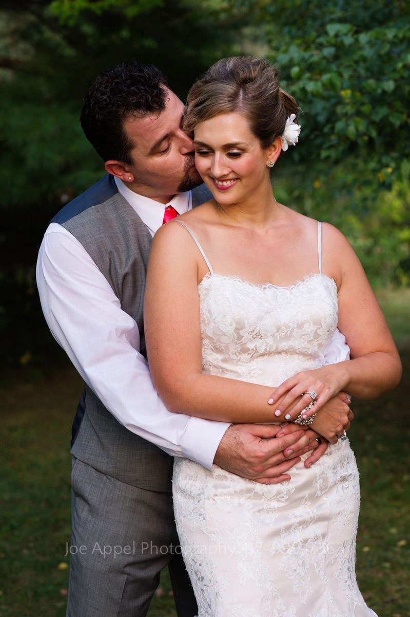 Bride smiles as groom wraps his arms around her and kisses her cheek.