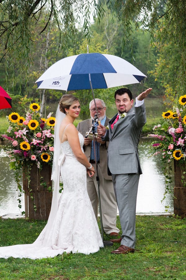 A couple stands beneath an umbrella as the groom sticks his hand out to test the rain during their wedding ceremony.