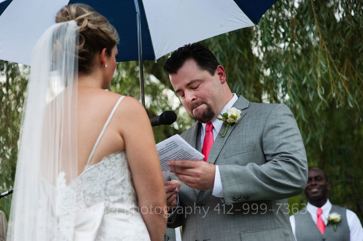 A groom fights back tears as he says his vows to his bride as they stand under an umbrella.