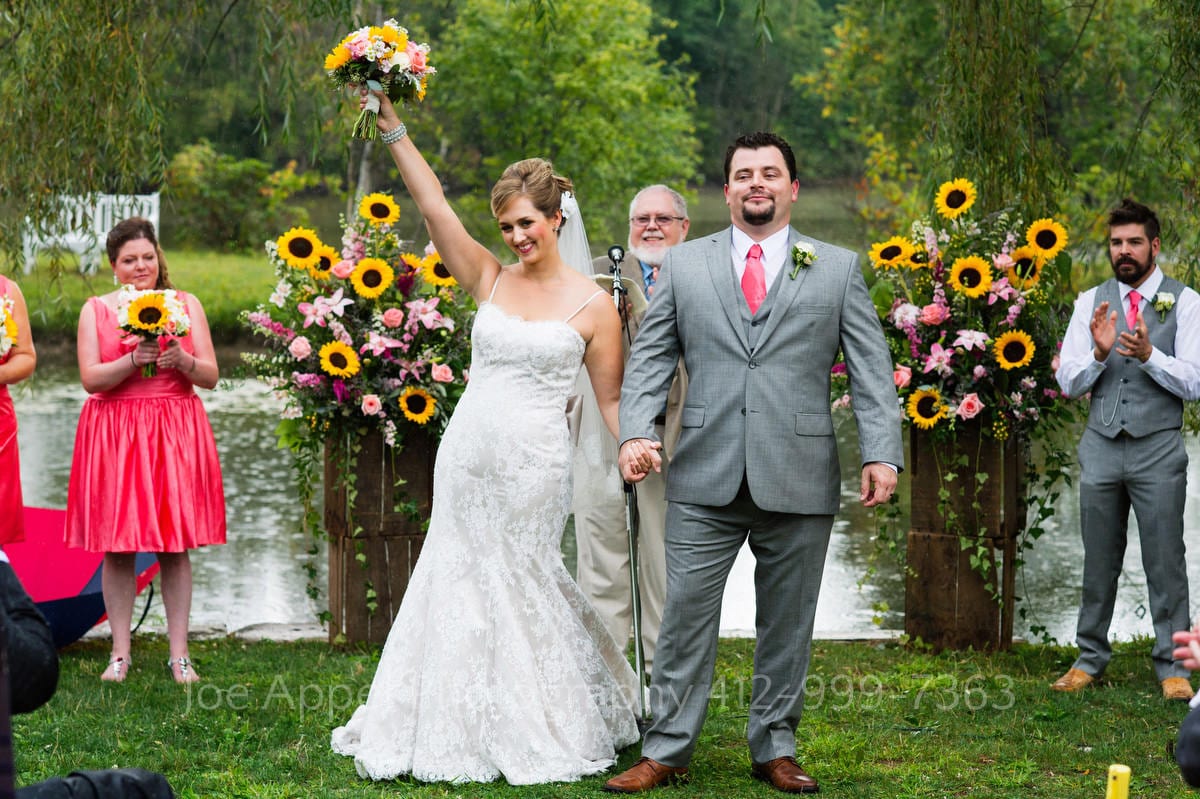 A bride raises her hand in celebration at the end of their wedding ceremony. The background is filled with bright yellow sunflowers.
