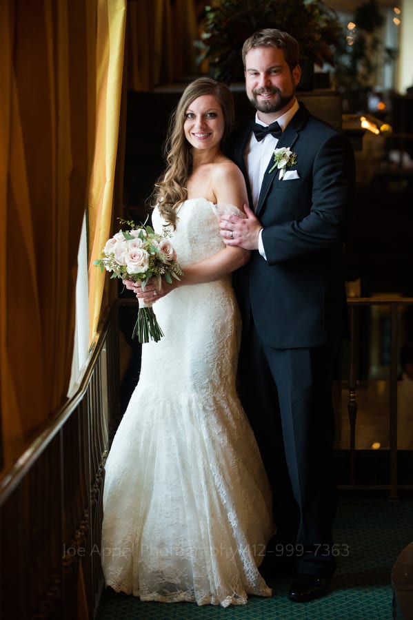 A handsome young married couple stands close to one another while facing a large window in the lobby of the William Penn Hotel in downtown Pittsburgh.
