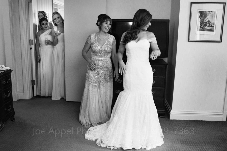 A bride puts on her dress with the help of her mother as her friends watch from the doorway to the hotel suite.
