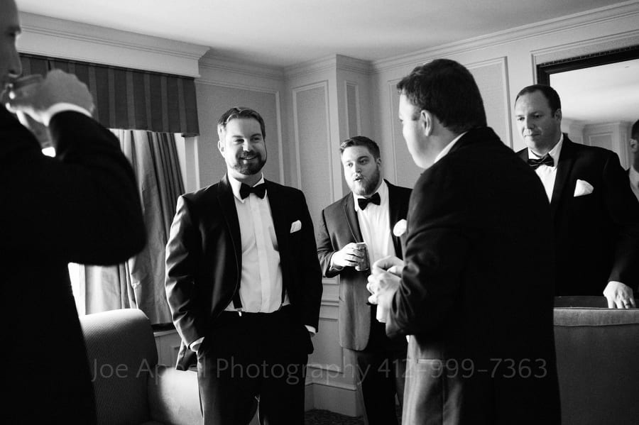A groom laughs with his groomsmen as they stand about a hotel room wearing tuxedos.