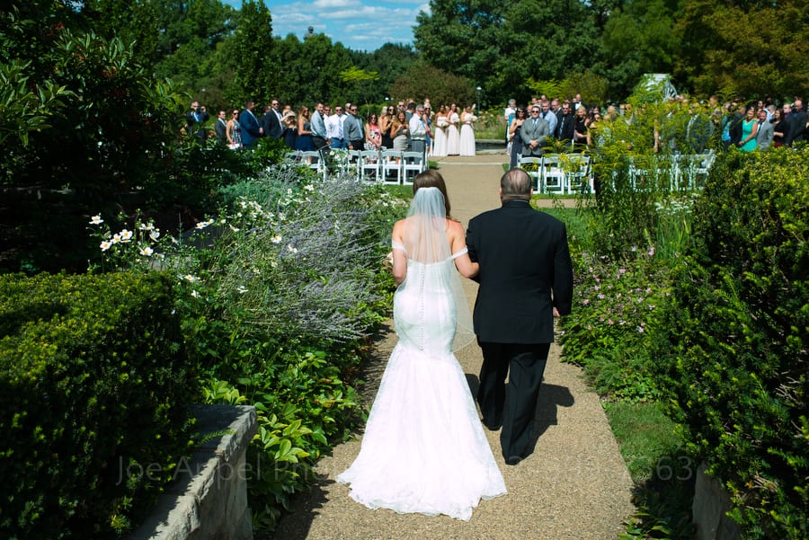 A bride and her father walk down a path towards the waiting wedding party outside at Phipps Conservatory.