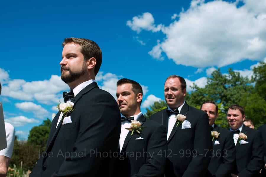 A groom waits for his bride under blue skies and puffy clouds.