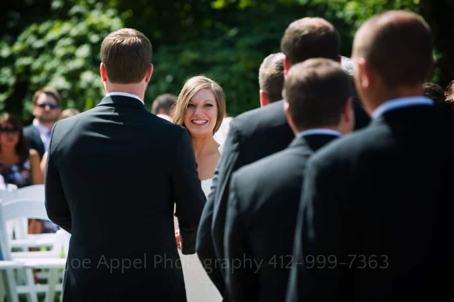 A bride's smiling face is seen through a gap between the groom and his groomsmen.