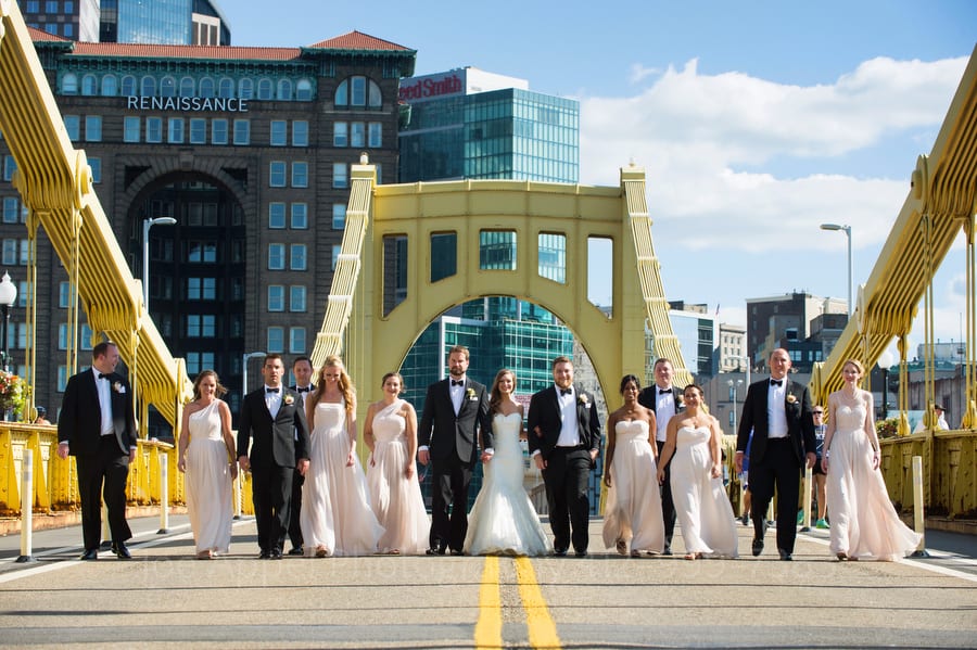 A large wedding party holds hands as they walk across both lanes of the Roberto Clemente bridge in downtown Pittsburgh.