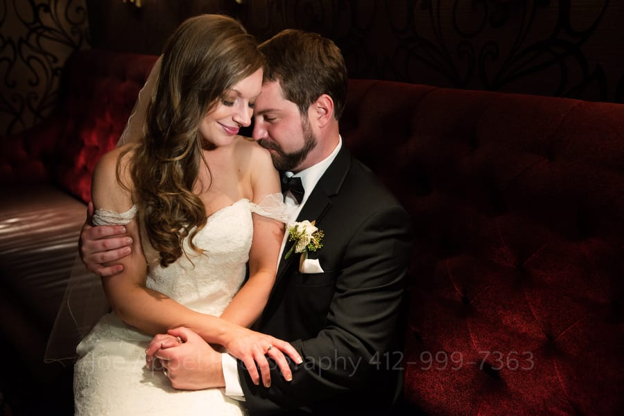 A bride and groom sit on a red velvet settee in the Speakeasy at their Omni William Penn wedding
