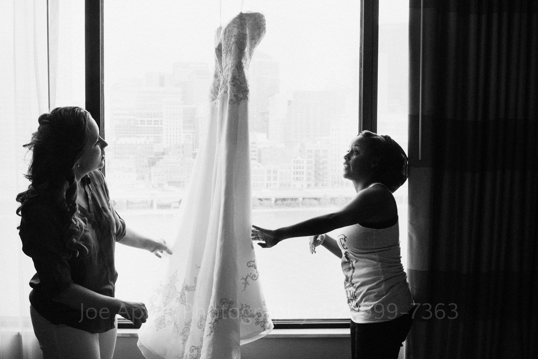 A bride and bridesmaid smooth out the wrinkles on a wedding dress hanging in the window of a hotel room.