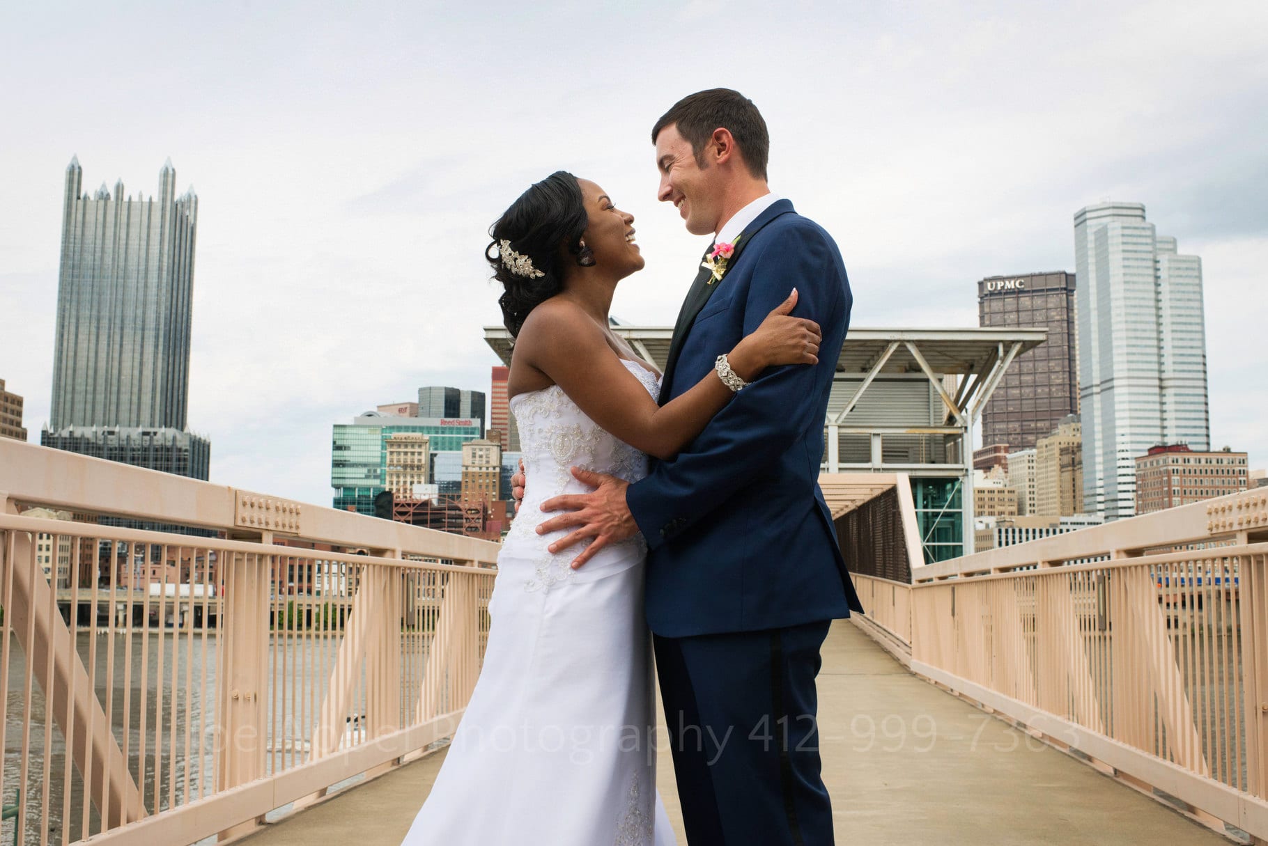 Bride and groom embrace on an elevated walkway at Station Square with the skyline of downtown Pittsburgh behind them.