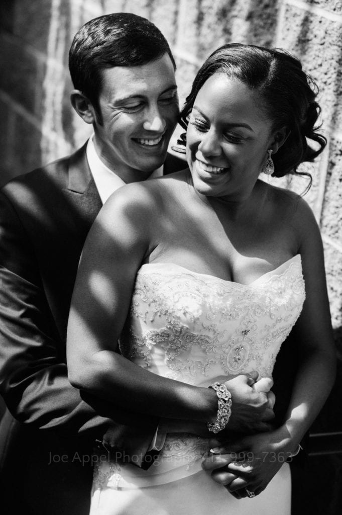 Groom pulls his bride into his chest as they smile in dappled sunlight.