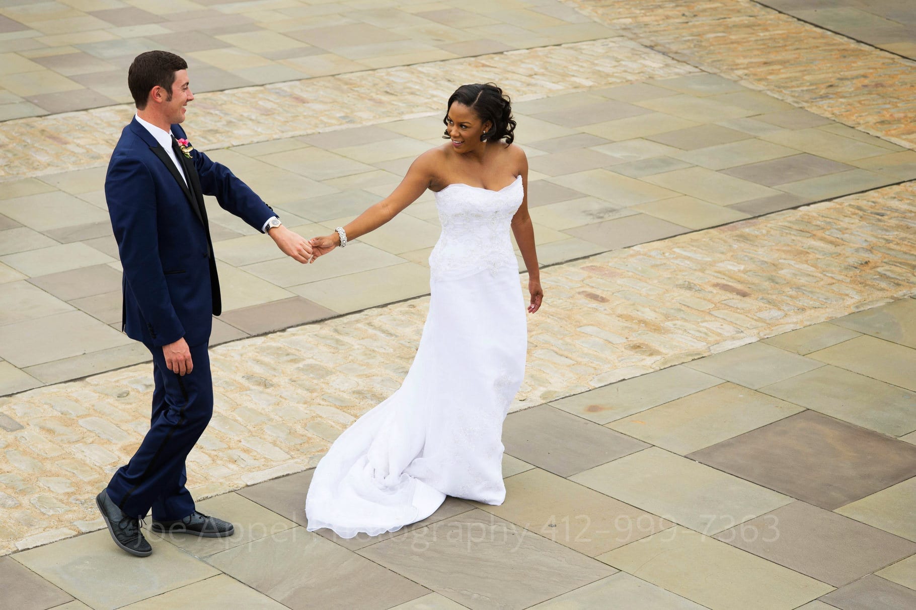 A bride and groom hold hands as they walk on the stone plaza next to the fountain in Point State Park.