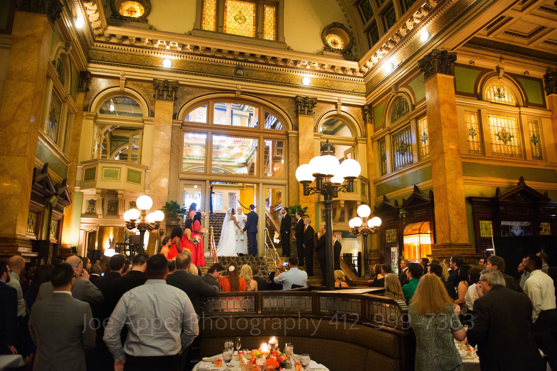 Guests stand on the ground floor of a large, golden-hued, ornate room as the bride and groom are married on the stairway above them. A classic scene during a Grand Concourse Wedding.