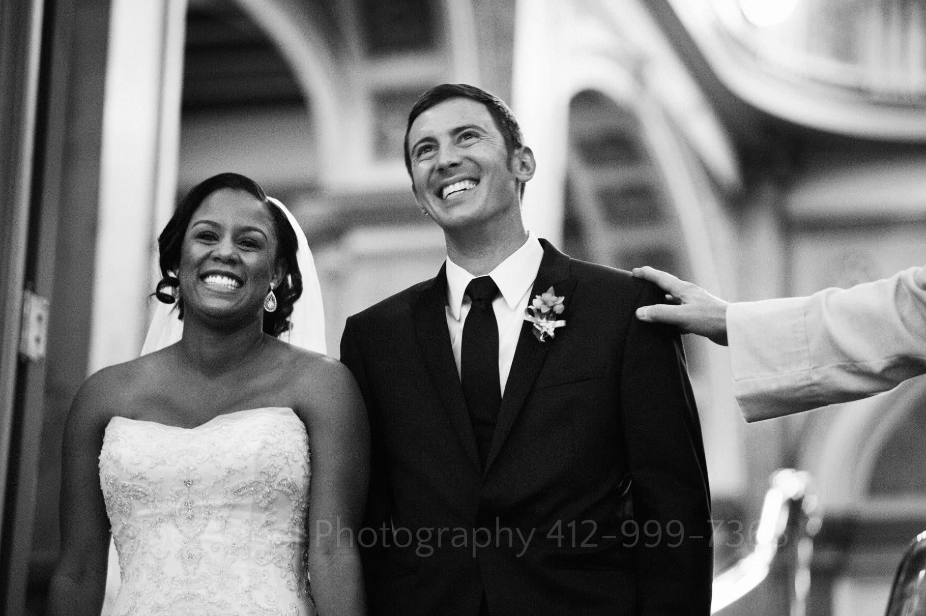 Bride and groom smile as they turn to look at their guests from atop a grand staircase. The reverend's hand is resting on the groom's shoulder.