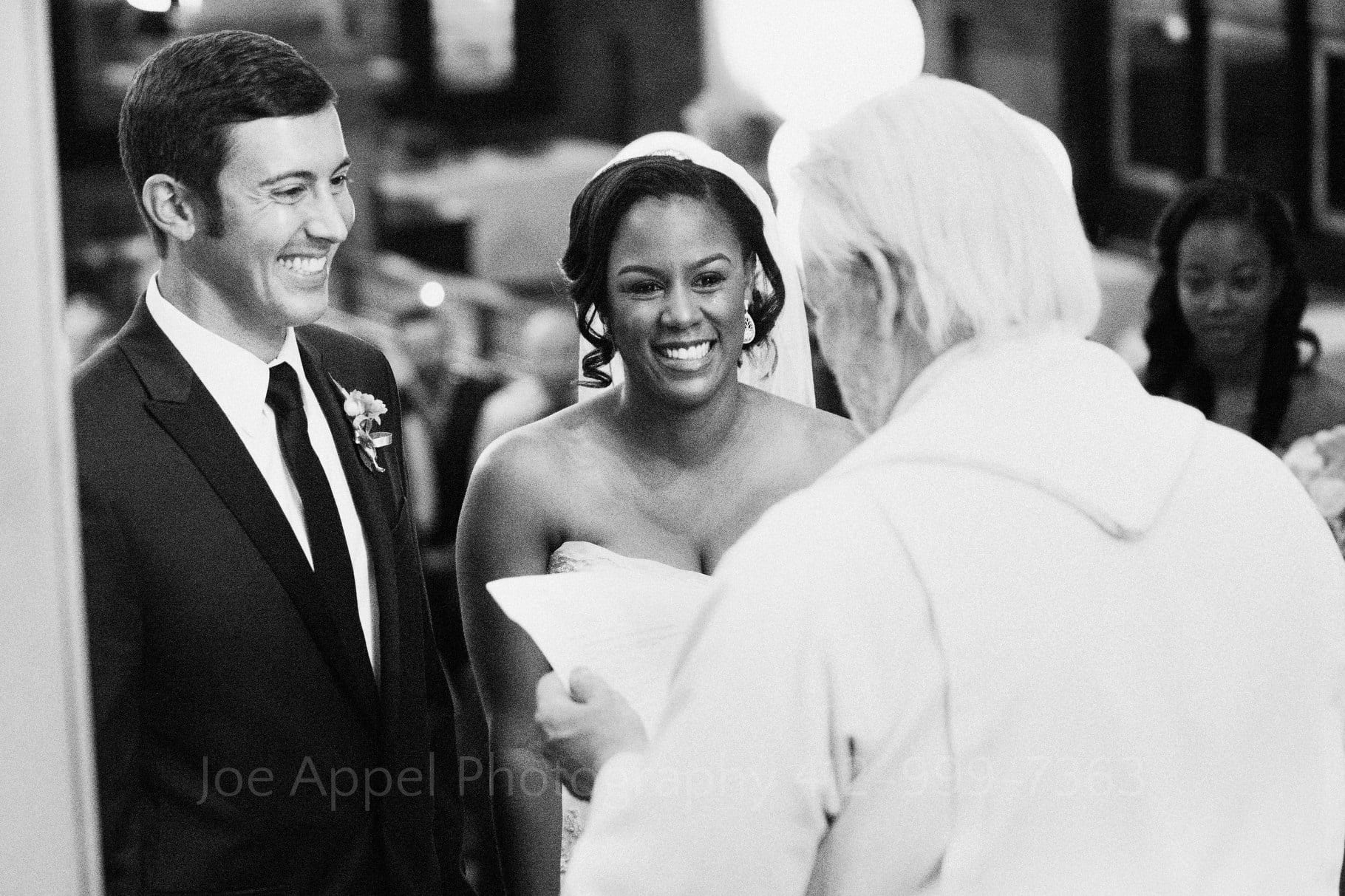 A bride and groom smile as they face their white-robed, and white-haired reverend during their Grand Concourse Wedding.