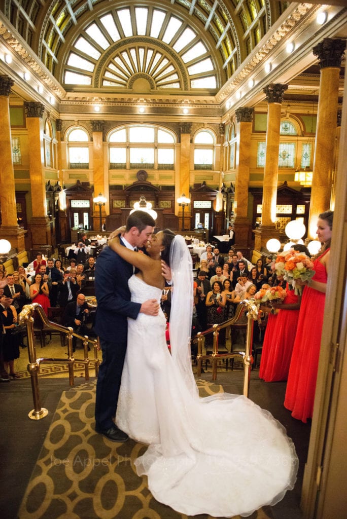 A bride and groom exchange a kiss atop a staircase as their guests applaud. The room is lit from above them by a large, flower-shaped stained glass window.