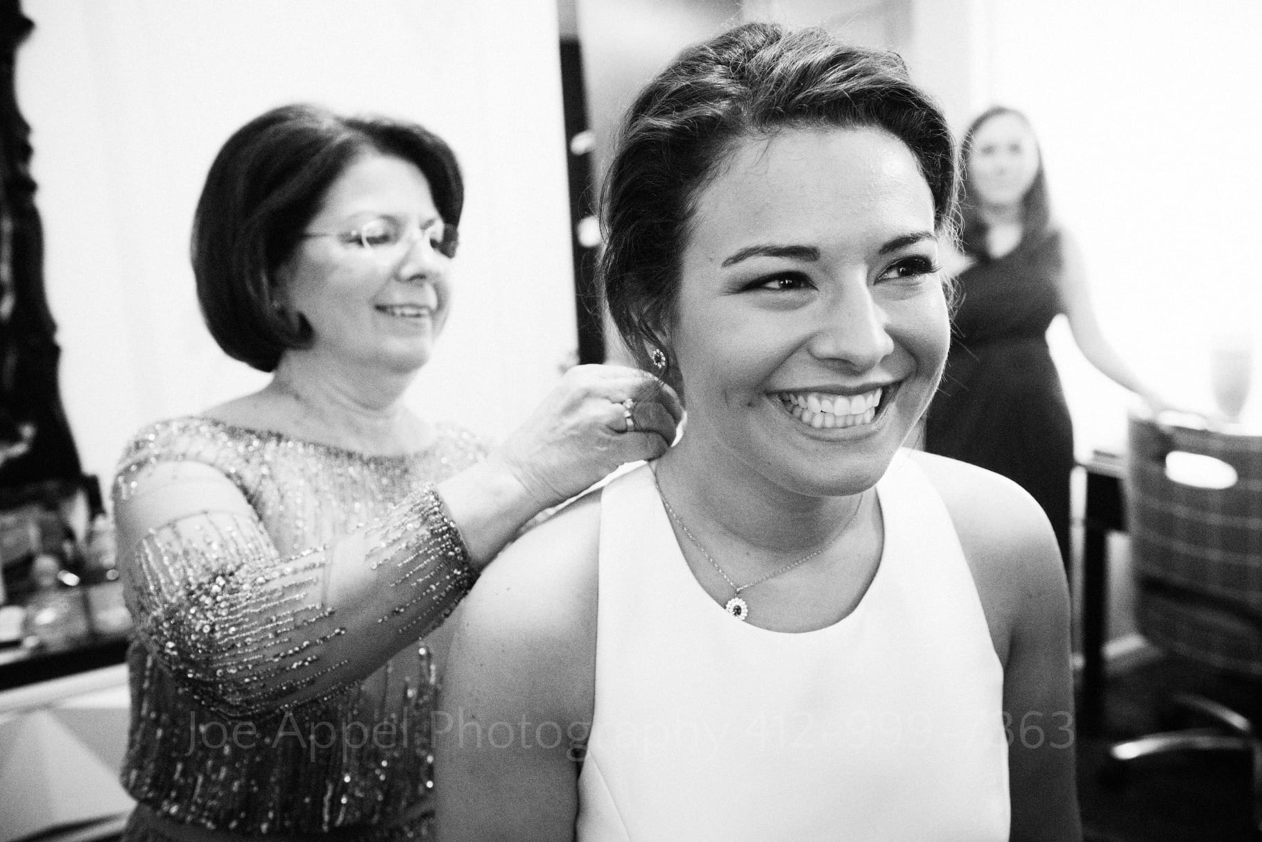 Black and white photo of a bride smiling in the foreground as her mom clasps a necklace around her neck.