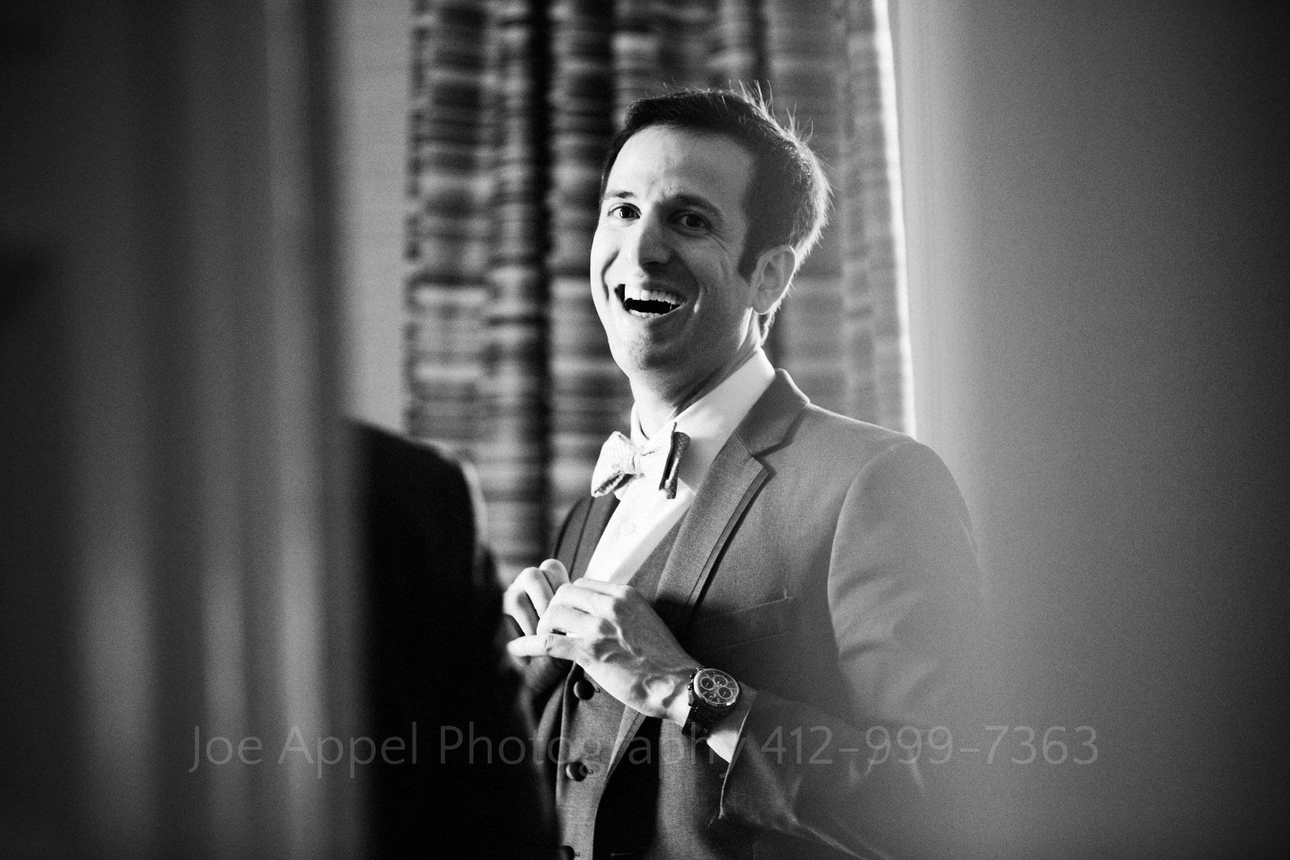 A young groom laughs as he buttons the shirt to his tuxedo in a room at the Duquesne Club.