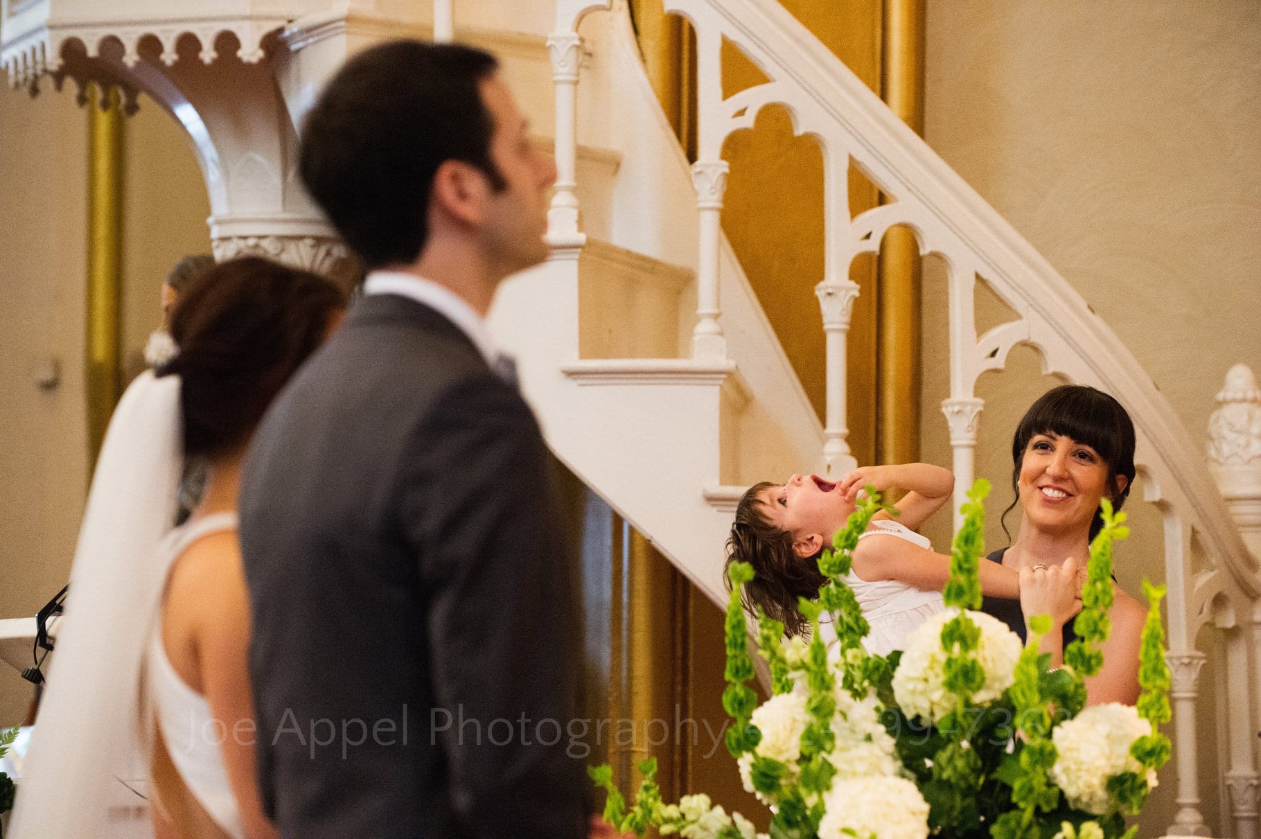 A bride and groom stand in the foreground while a bridesmaid holds her daughter in the background. The daughter lays back and looks at the ceiling with her mouth open.