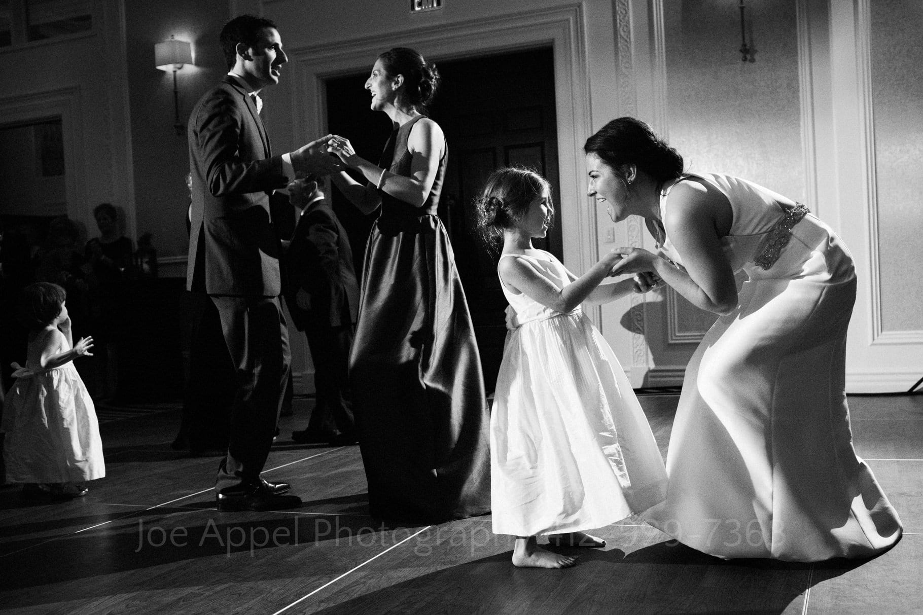 A bride bends down as she dances with a little girl. The groom dances with his sister behind them.