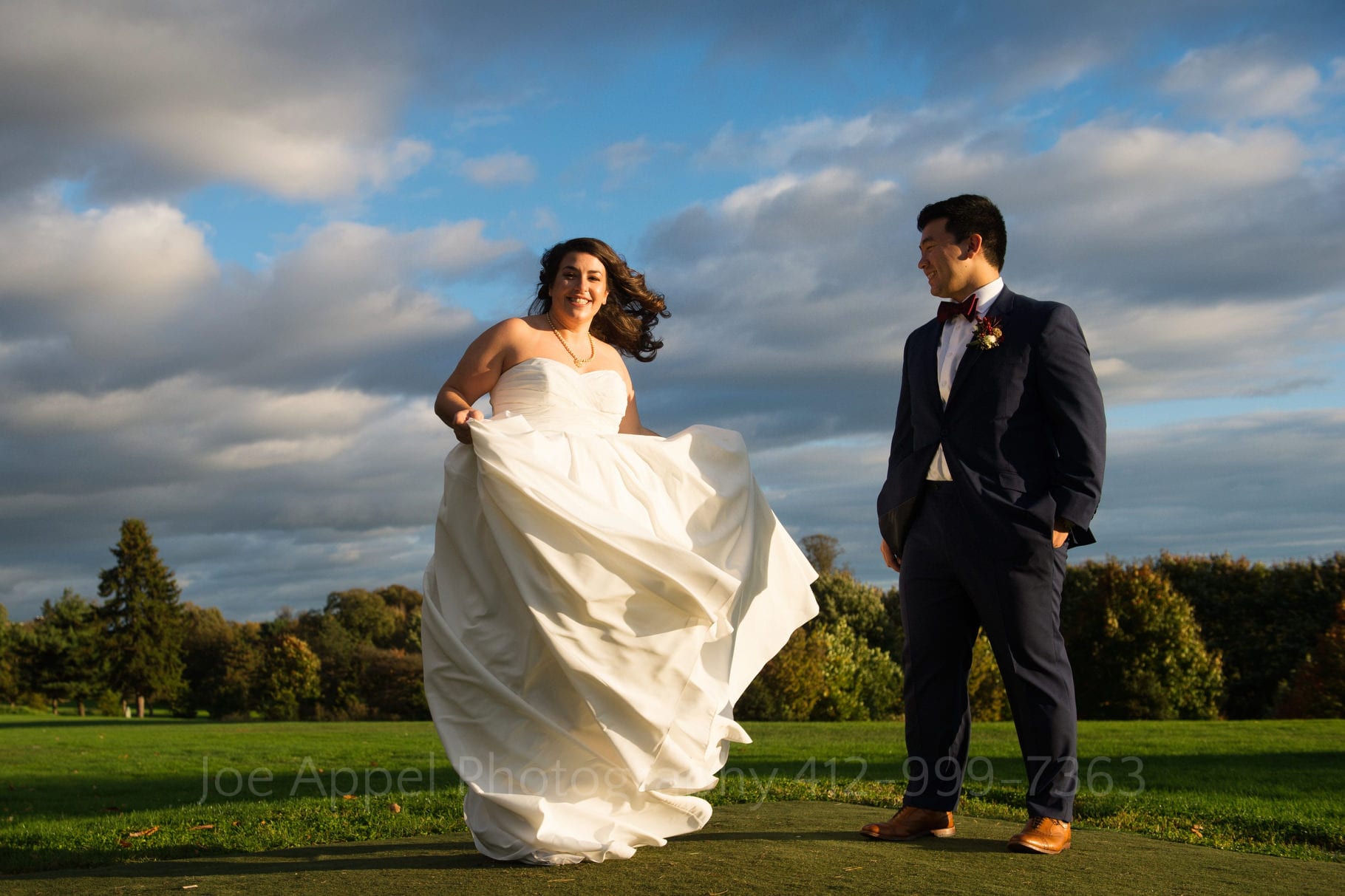 A bride and groom stand in the sunlight with puffy white clouds in the blue sky above them.