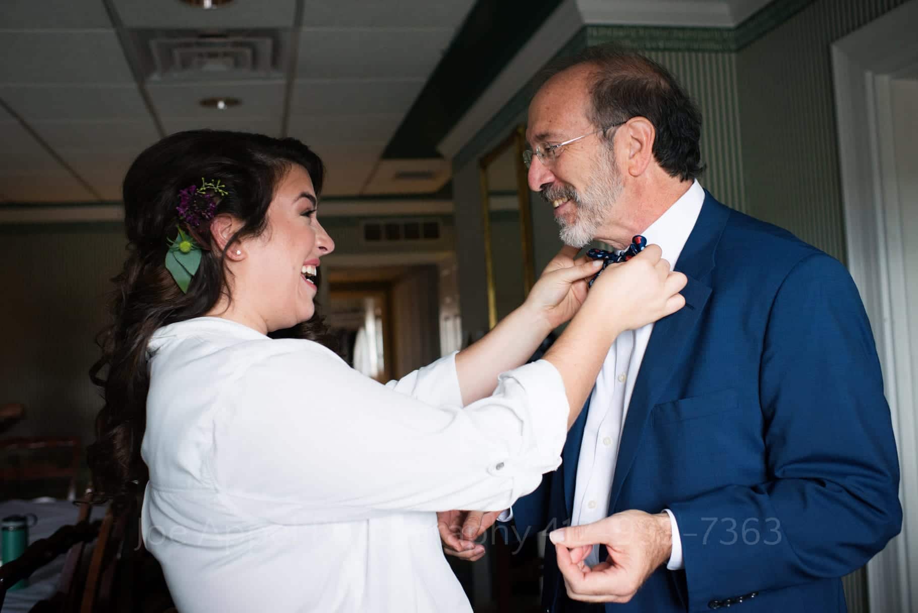 A young woman wearing a white bathrobe adjusts her father's bow tie as they smile at one another.