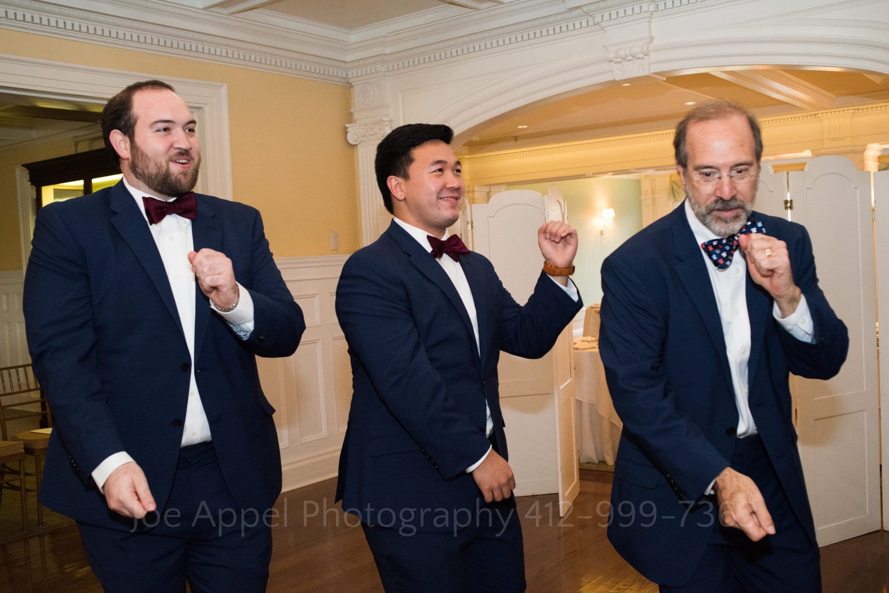 Three men wearing blue suits and bow ties practice their dance moves.