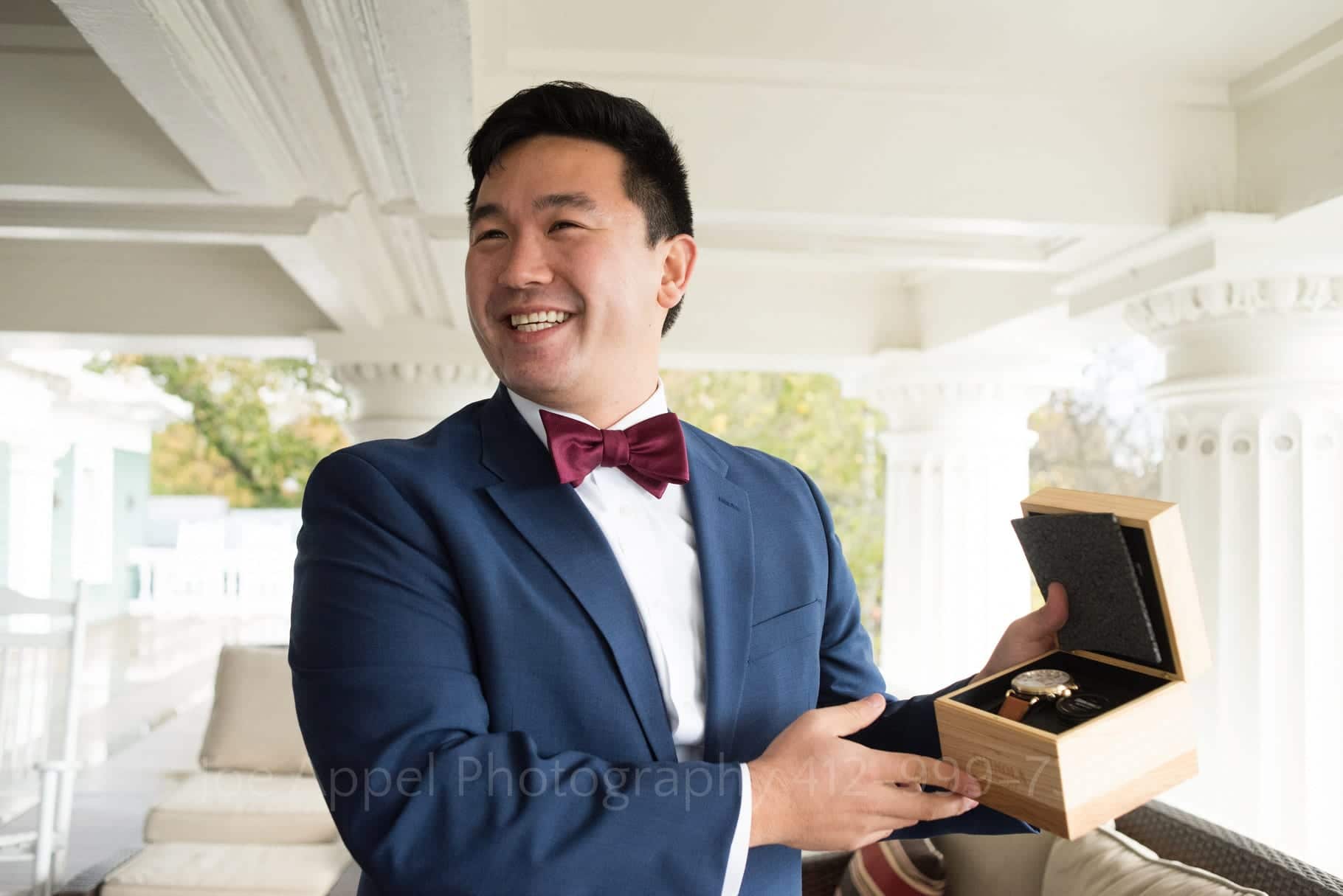 A groom wearing a blue suit and a pink bow tie smiles as he shows a box with a watch from Shinola that his bride gave him