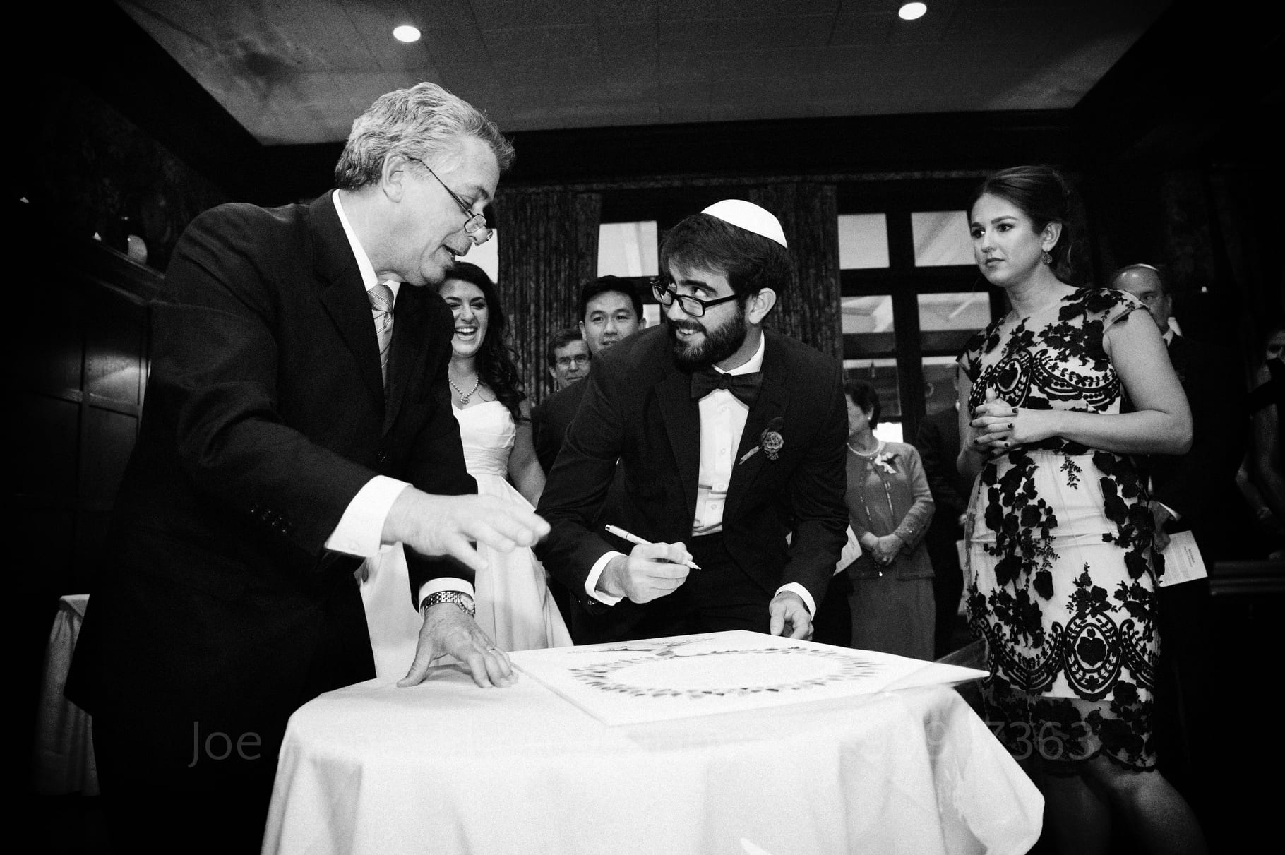 witness smiles as he looks at a rabbi who is giving him instruction on where to sign the ketubah during a wedding at the Pittsburgh Golf Club