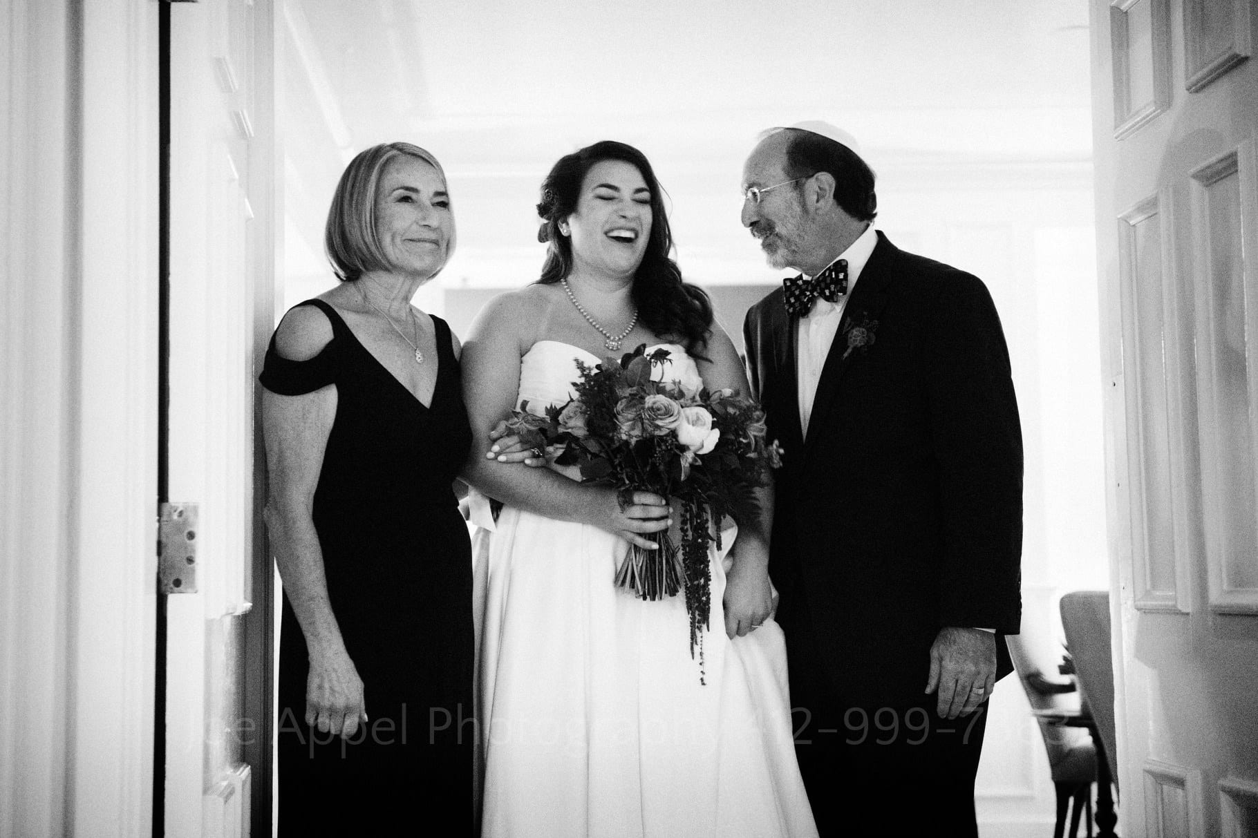 Bride smiles as she stands between her mother and father as they prepare to walk down the aisle at her wedding.