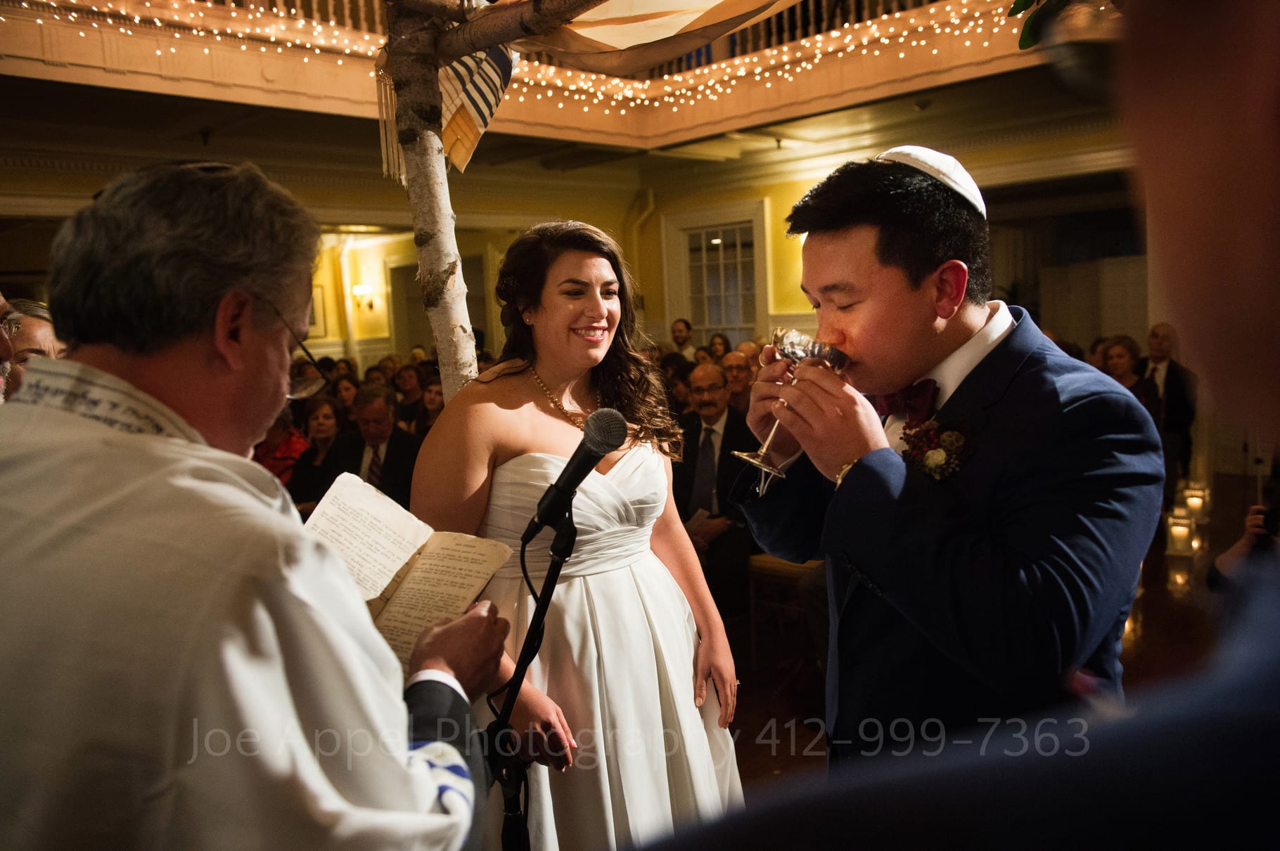 Groom drinks wine from a kiddush cup while his bride looks on smiling