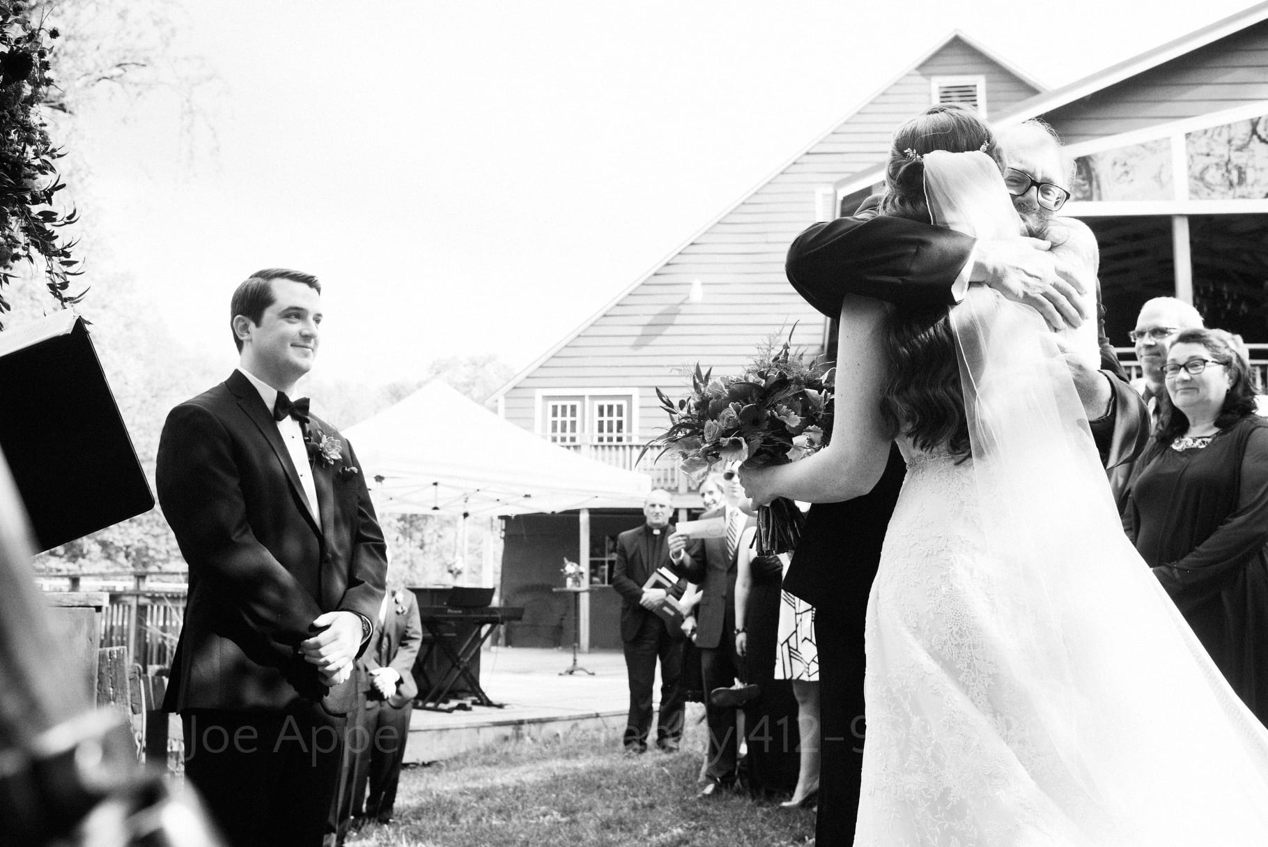 a groom smiles as he watches his bride and her father embrace at their wedding during their Chanteclaire Farm Wedding.