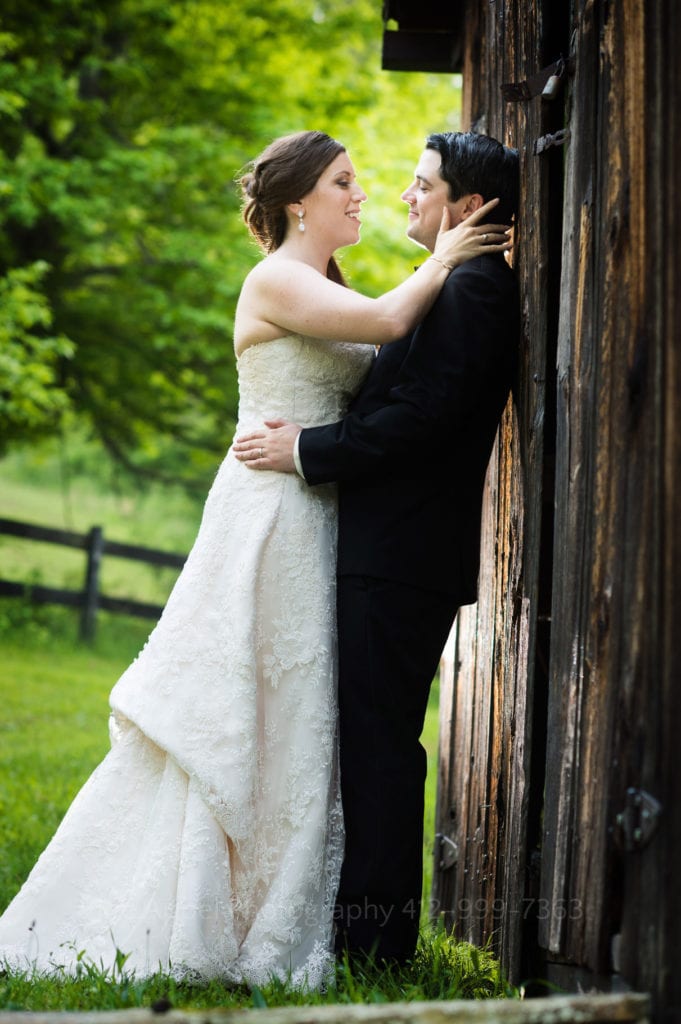 full length photo groom leans against a barn door while his bride caresses his cheek.