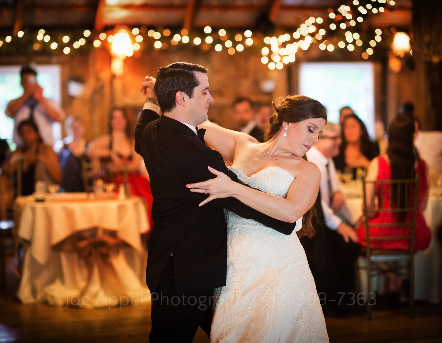 first dance bride looks away as she waltzes beneath twinkle lights inside of a barn