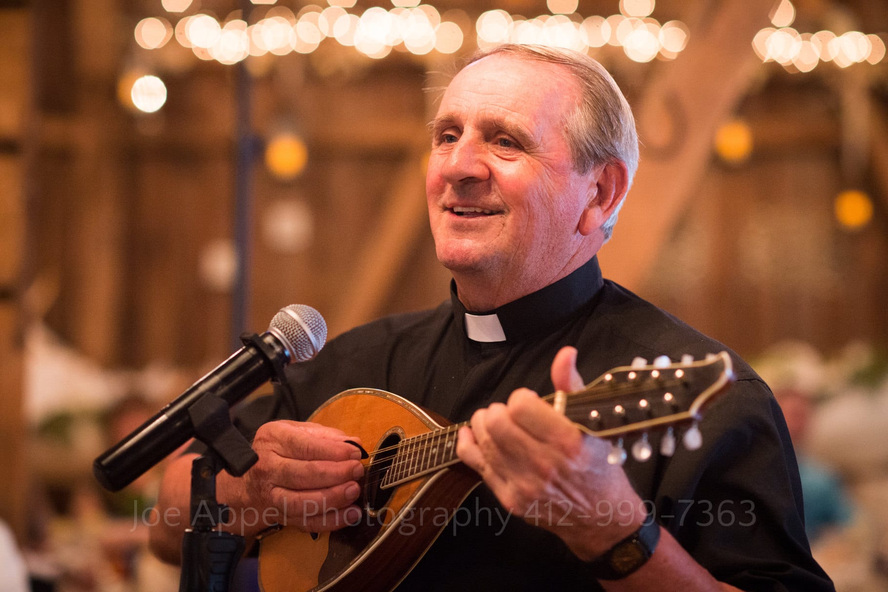 a priest sings as he plays a mandoline beneath twinkle lights.