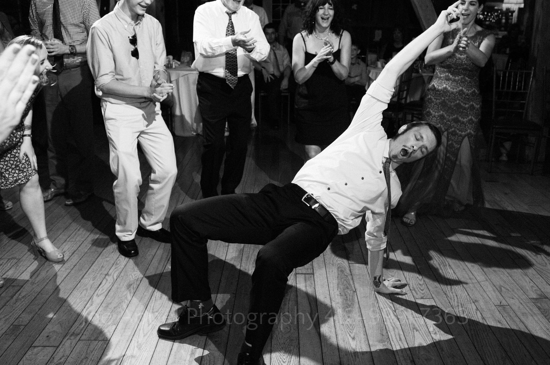 man bends over and touches the dance floor with one arm while the other arm is raised in the air. A group of guests stand around him during their Chanteclaire Farm Wedding.