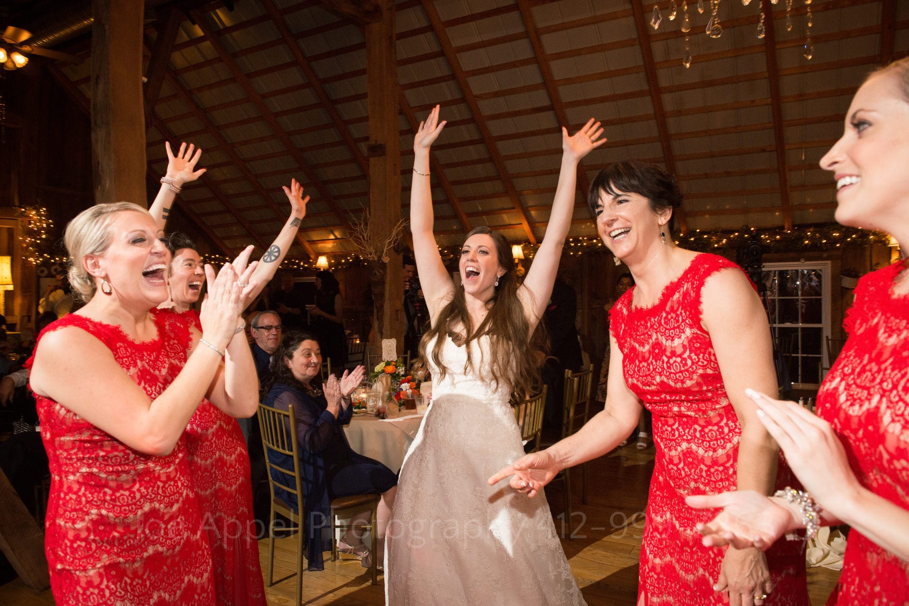 bride raises her hands in the air and smiles as she dances with her bridesmaids wearing red dresses during their Chanteclaire Farm Wedding.