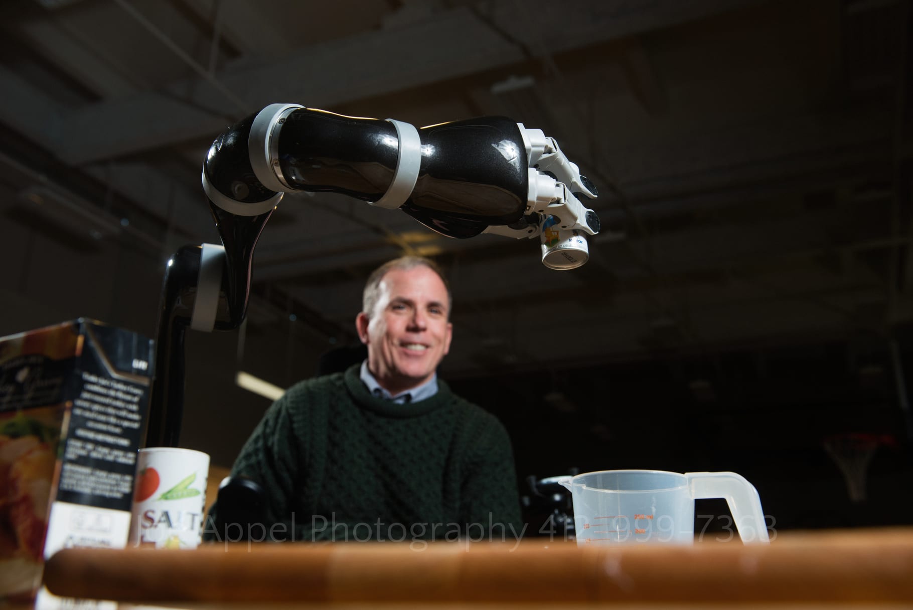 a man smiles as he manipulates a robotic arm to pour salt into a measuring cup by Pittsburgh Editorial Photographers Joe Appel Photography.