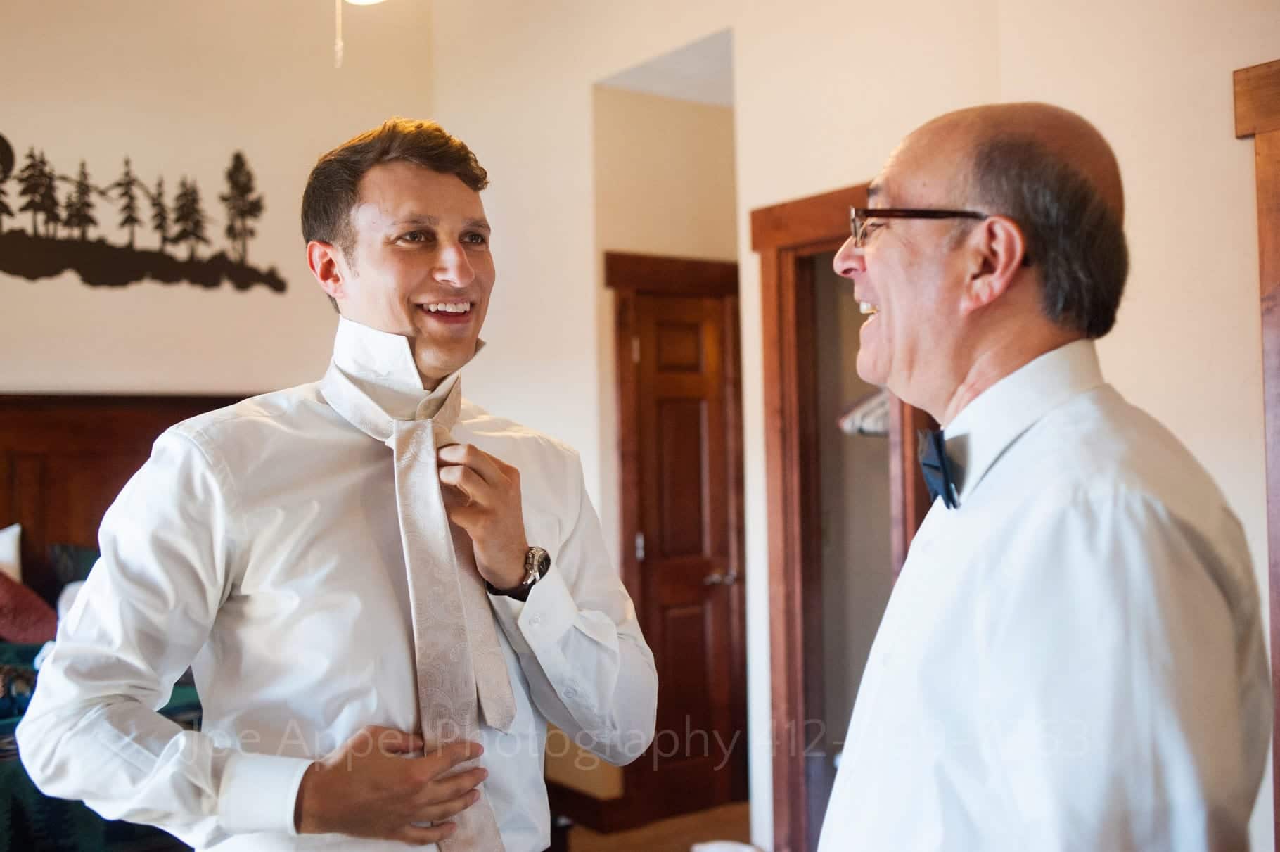 A young groom faces his father and smiles while putting on a pink tie before his wedding.