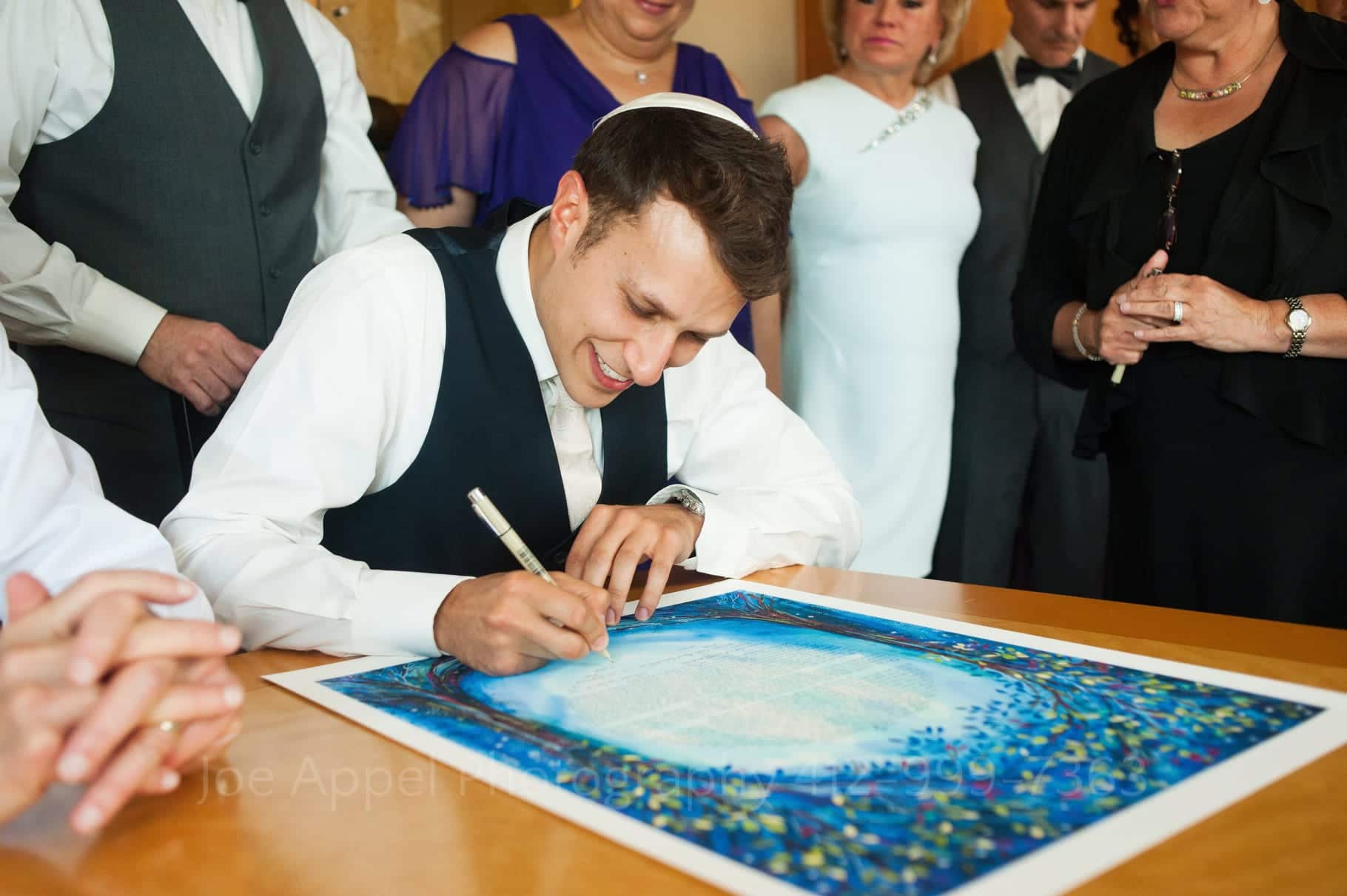A groom signs a blue ketubah on a brown table as family members gather around to witness.