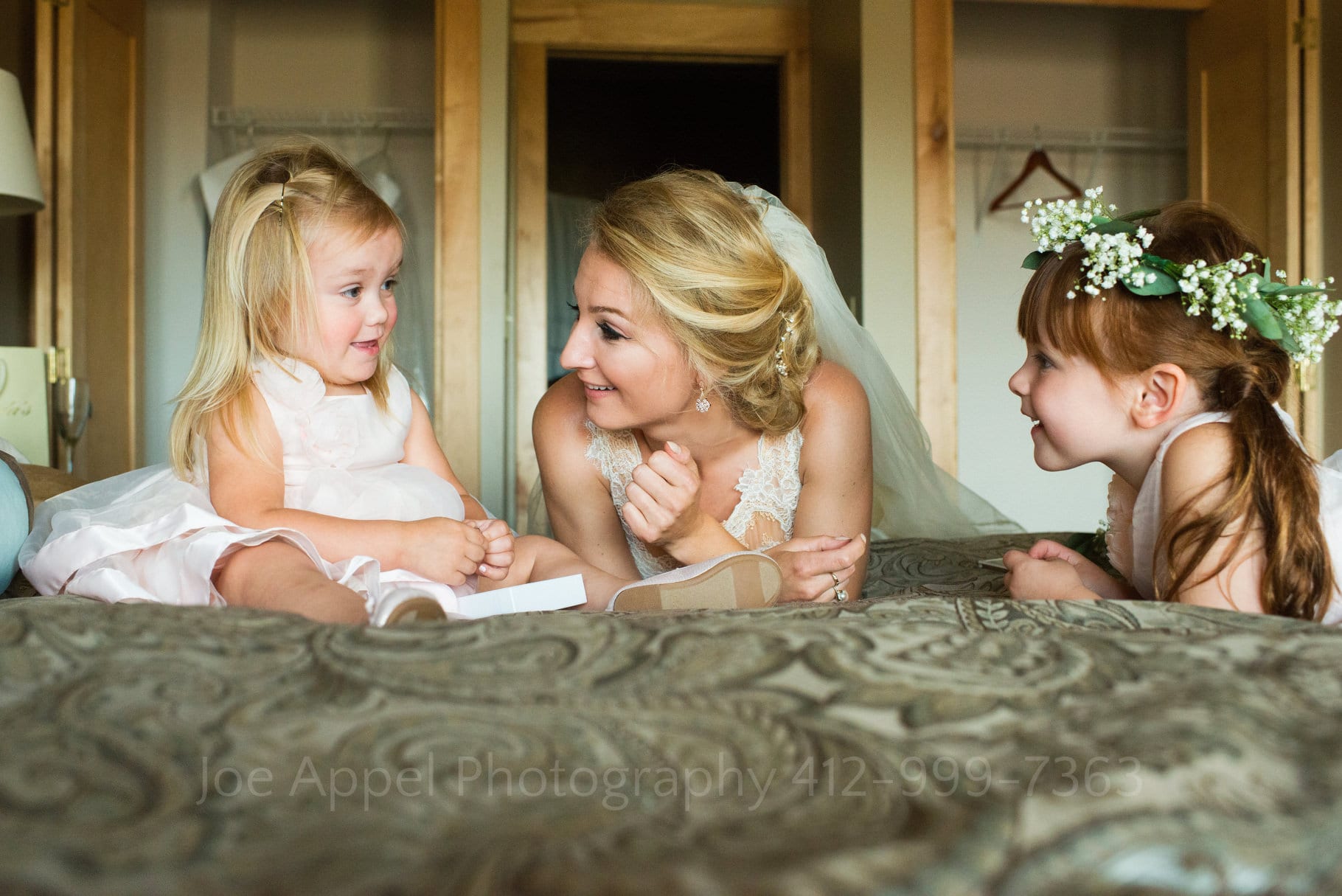 A bride lays on a bed between two little flower girls as they smile and talk.